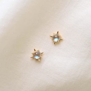 May include: A pair of gold stud earrings featuring a round, iridescent moonstone cabochon set in a four-pointed star frame. The stones display a soft blue glow against the polished gold metal, creating a celestial design for these small earrings.