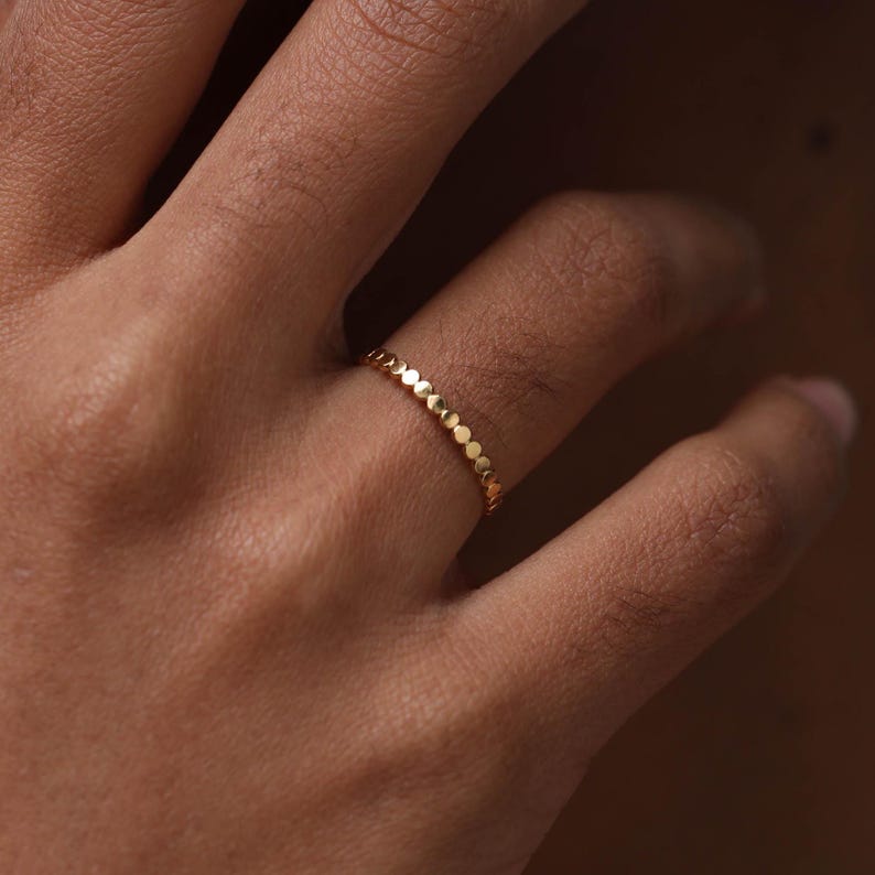 May include: A thin gold ring featuring a continuous band of small, circular, metallic discs linked together. The ring has a polished, reflective surface and a uniform, repeating pattern of round elements that encircle the entire circumference.