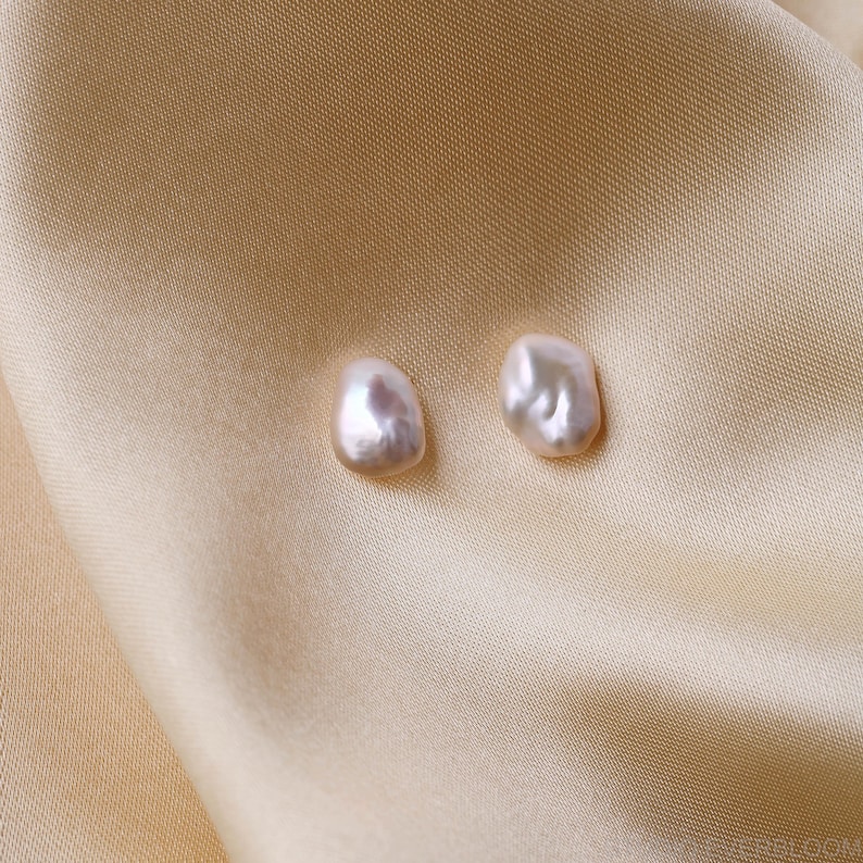 Pu&ograve; includere: Due orecchini di perle irregolari, bianco sporco. Le perle hanno una lucentezza lucida e una sfumatura leggermente rosata. Sono presentati su uno sfondo di tessuto beige satinato, creando un'estetica morbida ed elegante.