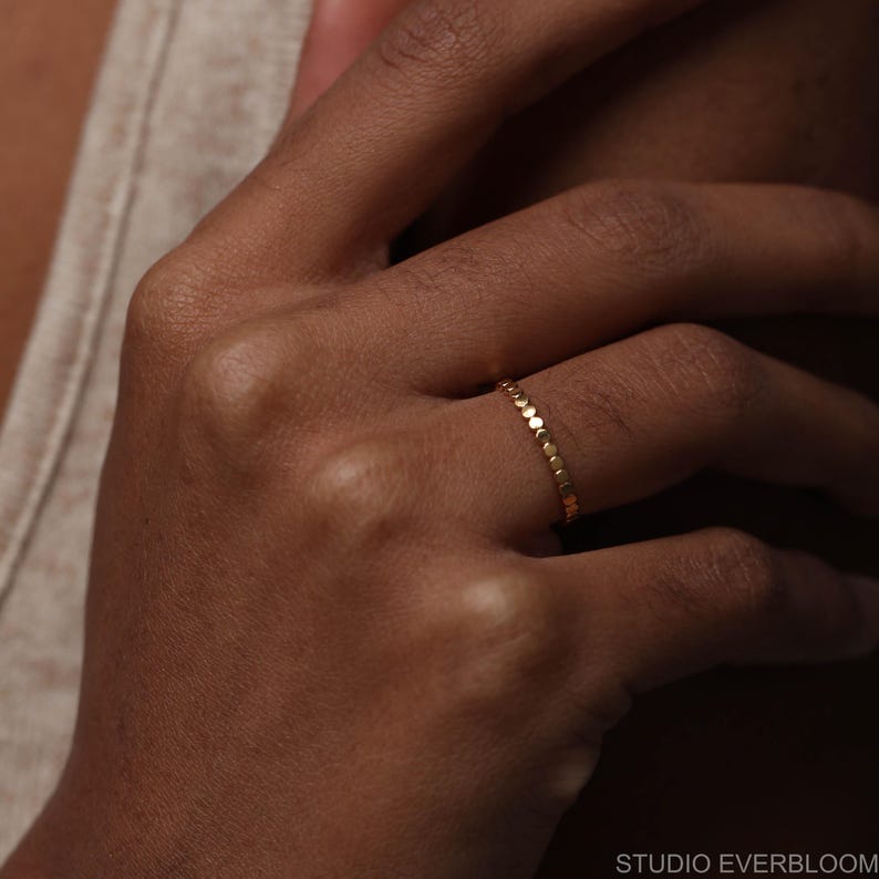 May include: A thin gold ring featuring a continuous band of small, circular, flat metal discs linked together. The ring has a polished, reflective surface and a uniform, delicate design that encircles the finger. The text STUDIO EVERBLOOM is visible.