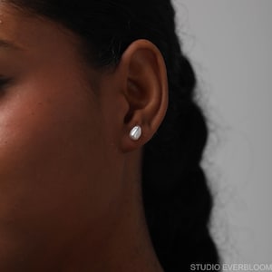 Pu&ograve; includere: Primo piano di un orecchio con un singolo orecchino a perno di perla bianca. L'orecchino, di forma ovale, &egrave; posizionato sul lobo dell'orecchio. Lo sfondo &egrave; di un colore chiaro e neutro.