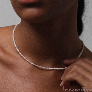 Può includere: Una delicata collana di perle bianche. La collana è composta da piccole perle rotonde infilate insieme, creando un design semplice ma elegante. La collana è indossata intorno al collo e l'immagine è un primo piano, che evidenzia i dettagli della collana.