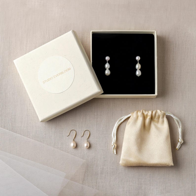 Op de afbeelding: Een paar oorbellen met elk twee witte parels, gepresenteerd in een vierkante gebroken witte doos met de tekst STUDIO EVERBLOOM op het deksel. Een tweede paar oorbellen en een klein beige trekkoordzakje zijn in de set inbegrepen.