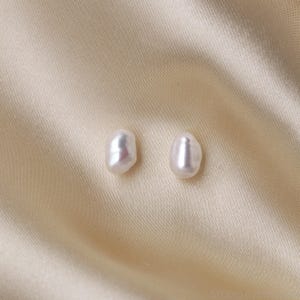 Pu&ograve; includere: Due orecchini di perle bianche di forma irregolare. Le perle hanno una lucentezza lucida e sono poste su uno sfondo di tessuto di raso beige chiaro. Un classico articolo di gioielleria.