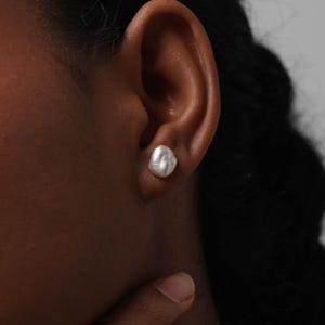 May include: A close-up of a person's ear with a white, irregular-shaped pearl stud earring. The earring is set against the skin, with the person's dark hair and skin visible. The earring is the focal point.