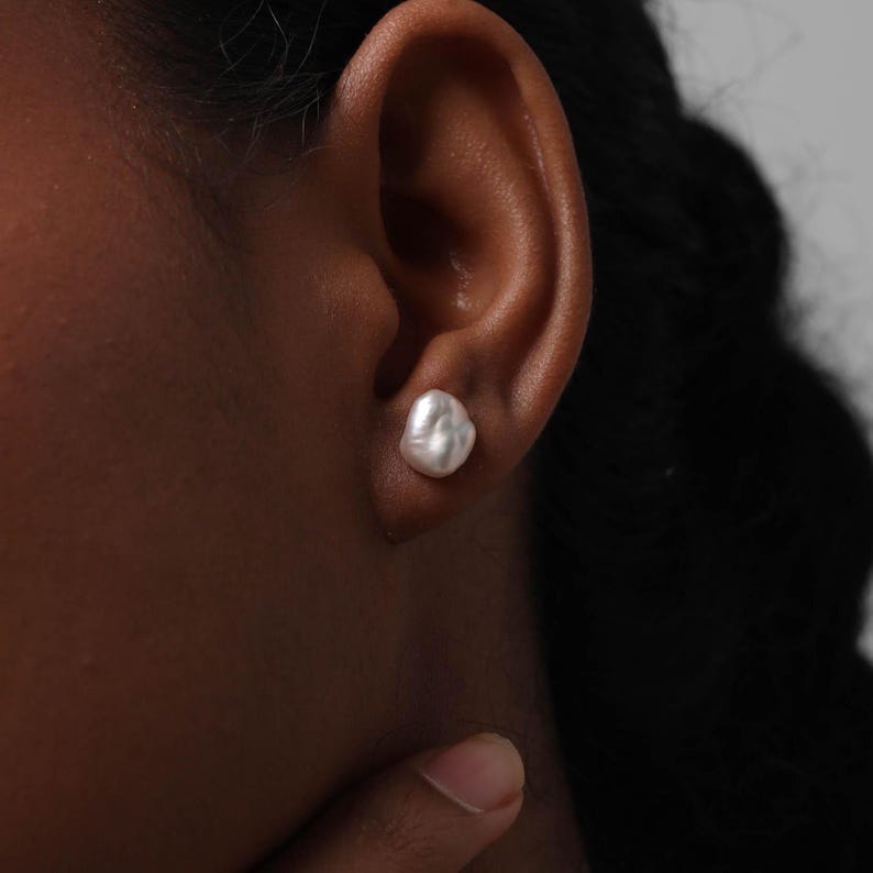 May include: A close-up of a person's ear with a white, irregular-shaped pearl stud earring. The earring is set against the skin, with the person's dark hair and skin visible. The earring is the focal point.