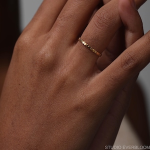 May include: A thin gold ring featuring a continuous band of small, circular, flat-topped metal discs. The ring has a polished finish and a uniform, repeating pattern of connected round elements that encircle the entire circumference of the band.