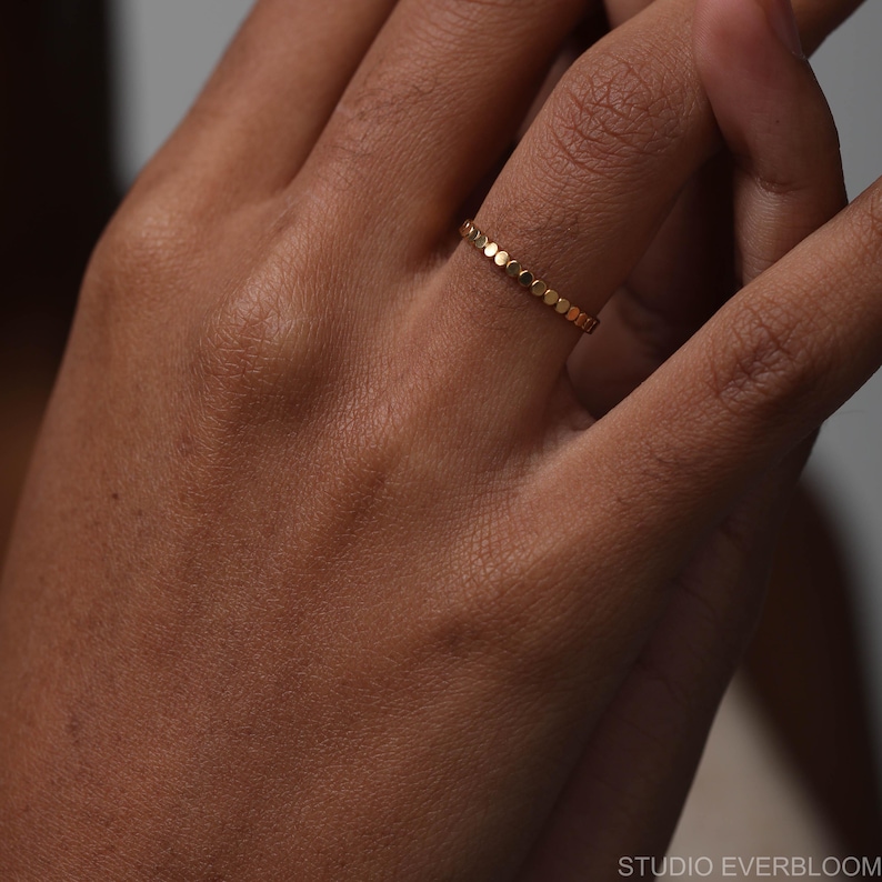 May include: A thin gold ring featuring a continuous band of small, circular, flat-topped metal discs. The ring has a polished finish and a uniform, repeating pattern of connected round elements that encircle the entire circumference of the band.