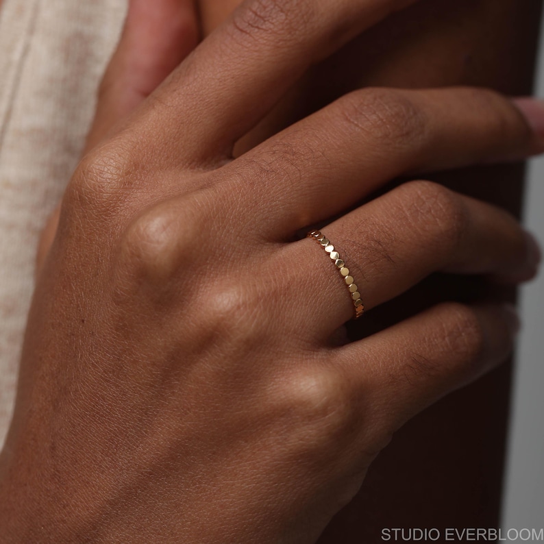 May include: A thin gold ring featuring a continuous band of small, circular, flat metal discs linked together. The ring has a polished, reflective finish and a delicate, uniform structure that encircles the finger, creating a textured appearance.