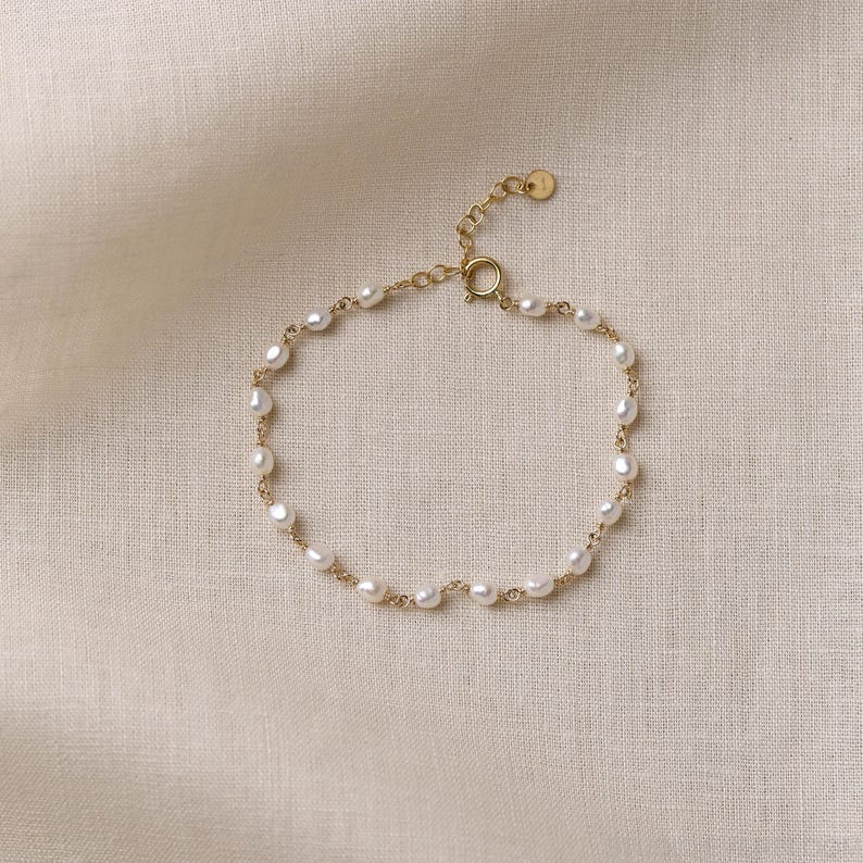 Op de afbeelding: Een delicate gouden kettingarmband met kleine, onregelmatig gevormde witte zoetwaterparels die gelijkmatig over de schakels zijn verdeeld. De armband is voorzien van een ronde veerringsluiting en een klein, rond gouden label bij de sluiting.