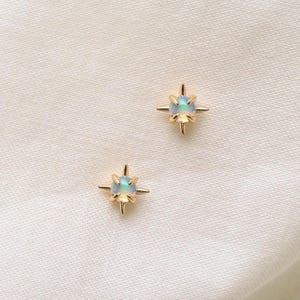 May include: A pair of gold stud earrings, each featuring a round, iridescent opal stone set in a four-prong starburst-shaped metal frame. The design highlights a central gemstone with a subtle play of colour, secured by pointed metal rays.