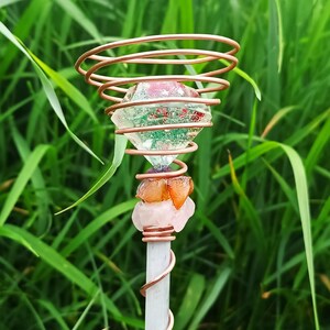 Può includere: Una bacchetta di cristallo fatta a mano con punta in quarzo trasparente, ornata con filo di rame e varie pietre preziose. La bacchetta presenta un grande cristallo sfaccettato racchiuso in spirali di rame, con pietre aggiuntive nei toni del rosa e dell'arancione. Sfondo di erba verde sfocata.