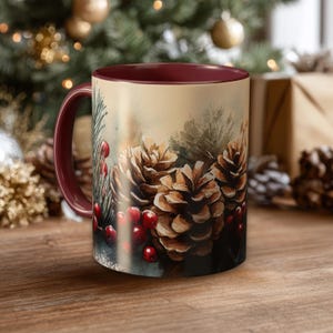 May include: A ceramic mug with a deep burgundy handle and interior rim. The exterior features a watercolor-style illustration of brown pinecones, green pine needles, and bright red berries against a soft, light-colored background.