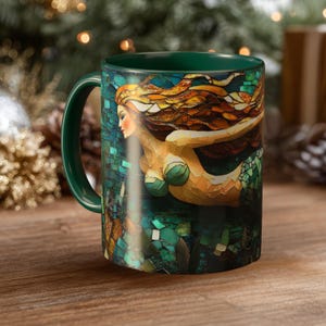 Puede incluir: Taza de café de cerámica con asa e interior verdes. La taza presenta una ilustración de estilo mosaico de una sirena con cabello castaño rojizo y un top de concha verde. La sirena está sobre un fondo verde azulado.