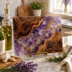 May include: A rectangular glass cutting board featuring a faux wood grain pattern on the left and right sides, separated by a central diagonal band of purple amethyst crystals with gold accents. The text Olivia's KITCHEN is printed in white.