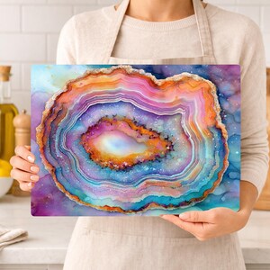 May include: A rectangular cutting board featuring a colorful geode design. The geode has layers of orange, pink, purple, and blue, with a white center and a sparkling, textured border. The background has a watercolor effect.