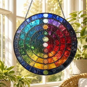 Op de afbeelding: Een ronde glas-in-lood zanger met een levendig regenboogspectrum in concentrische ringen. Een verticale kolom van zeven afzonderlijke ronde glasstukken in verschillende kleuren loopt door het midden, gevat in een zwart metalen frame.