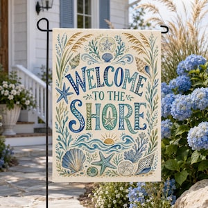 May include: A rectangular garden flag featuring the text "WELCOME TO THE SHORE" in blue and green lettering. The design includes illustrations of seashells, starfish, sand dollars, seaweed, and a rising sun, all set against a cream background.