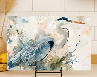 May include: A rectangular glass cutting board featuring a watercolor illustration of a great blue heron. The bird is depicted in shades of blue, gray, and white, set against a soft, abstract background of muted peach, green, and blue paint washes.
