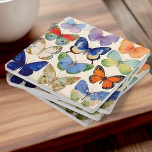 Puede incluir: Una pila de posavasos cuadrados con un estampado de acuarela de varias mariposas en tonos azul, naranja, morado, amarillo y verde sobre un fondo blanco roto. Las mariposas se muestran con alas y patrones detallados.
