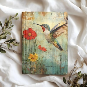 Può includere: Un diario rettangolare con un design di colibrì e fiori. La copertina presenta un colibrì colorato con le ali spiegate, fiori rossi, arancioni e gialli e uno sfondo strutturato e invecchiato. Il diario è appoggiato su un tessuto bianco.