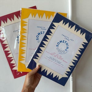May include: Three picture frames in red, yellow, and navy blue, each with a white zig-zag border. The frames are packaged in clear plastic. The text "Crafty Joy From Something Good Workshop" is printed on the front.