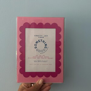 May include: A pink picture frame with a scalloped edge. The frame has a white interior with text that reads "Crafty Joy From Something Good Works". The frame is designed to hold A6 to A5 pictures or photo mounts.