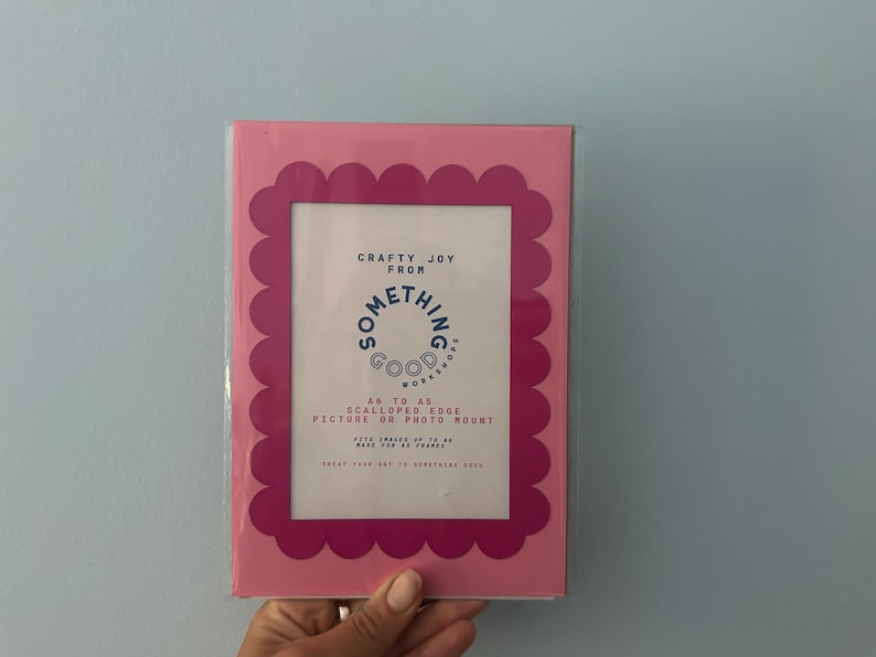 May include: A pink picture frame with a scalloped edge. The frame has a white interior with text that reads "Crafty Joy From Something Good Works". The frame is designed to hold A6 to A5 pictures or photo mounts.