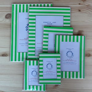 Könnte beinhalten: Eine Sammlung von grün-weiß gestreiften Bilderrahmen in verschiedenen Größen. Jeder Rahmen hat einen weißen Innenraum und trägt den Text "Crafty Joy From Something Good" und "Screenprinted Picture". Die Rahmen sind auf einer Holzoberfläche angeordnet.