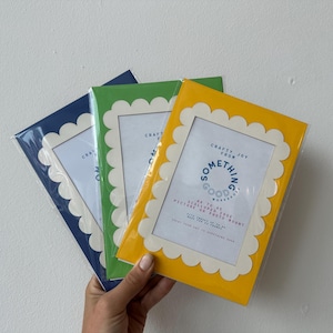 May include: Three picture frames in navy blue, green, and yellow, each with a scalloped edge and a white border. The yellow frame is held in a hand. Each frame has text that reads "Crafty Joy From Something Good Works".