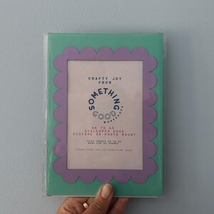 May include: A rectangular picture frame with a scalloped edge in a mint green and lavender color scheme. The frame has text that reads "Crafty Joy From Something Good Workers". It is designed to hold A6 sized pictures or photo mounts.