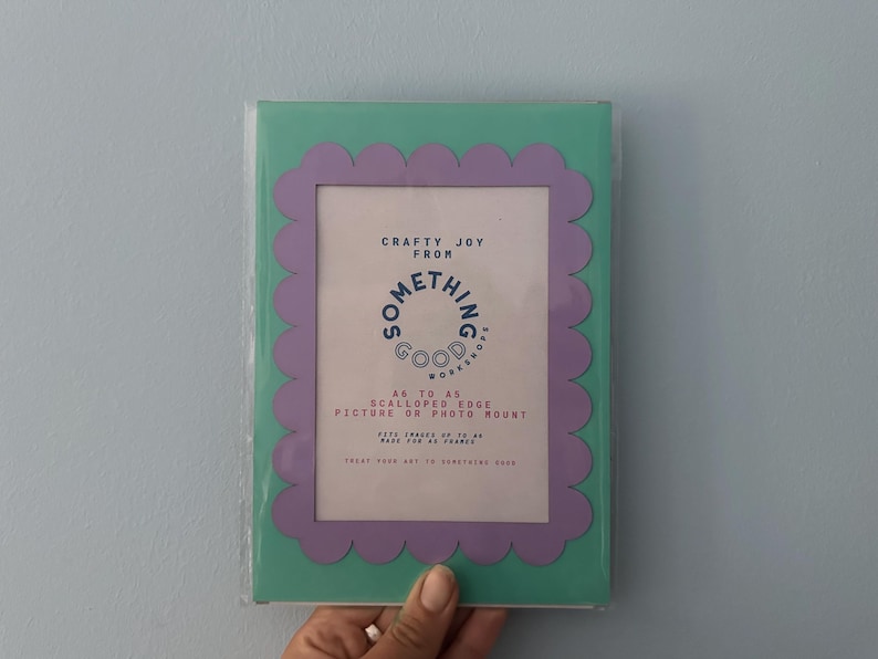 May include: A rectangular picture frame with a scalloped edge in a mint green and lavender color scheme. The frame has text that reads "Crafty Joy From Something Good Workers". It is designed to hold A6 sized pictures or photo mounts.