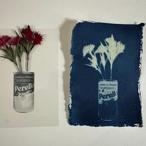 Op de afbeelding: Twee kunstwerken met bloemen in een Perelló conservenblik. Eén is een foto van rode bloemen. De andere is een cyanotypie-afdruk van witte bloemen op een donkerblauwe achtergrond.