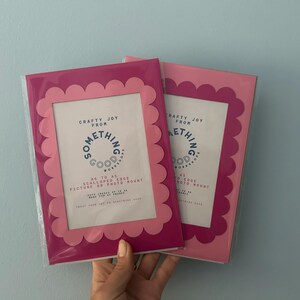 May include: Two pink picture frames with scalloped edges. The frames have a rectangular opening for photos and the text "Crafty Joy From Something Good Works" is printed on the front. The frames are packaged in clear plastic.