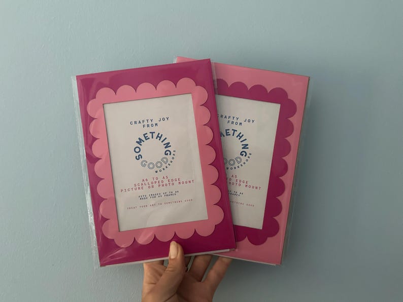 May include: Two pink picture frames with scalloped edges. The frames have a rectangular opening for photos and the text "Crafty Joy From Something Good Works" is printed on the front. The frames are packaged in clear plastic.