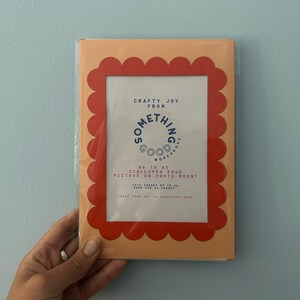 May include: A picture frame with a scalloped edge in a peach color, with a red border. The frame has the text "Crafty Joy From Something Good Workshop" and is designed for pictures or photo mounts.