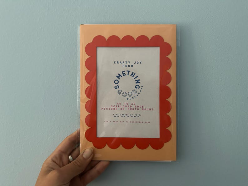 May include: A picture frame with a scalloped edge in a peach color, with a red border. The frame has the text "Crafty Joy From Something Good Workshop" and is designed for pictures or photo mounts.