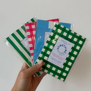 Könnte beinhalten: Eine Hand hält eine Sammlung farbenfroher Fotohalterungen. Die Halterungen weisen verschiedene Muster auf, darunter grün-weiße Streifen, rosa Vichy und grünes Vichy. Jede Halterung ist in klarem Kunststoff verpackt. Die grüne Vichy-Halterung trägt den Text "SOMETHING GOOD".