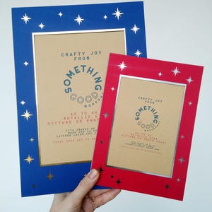 May include: Two picture frames, one blue and one red, with star cutouts and metallic silver inner frames. The blue frame has gold stars, and the red frame has silver stars. Text on the frames reads "Crafty Joy From Something Good Works."