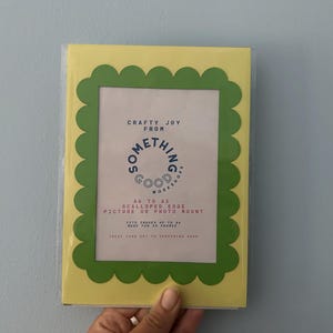May include: A yellow picture frame with a green scalloped edge. The frame has a white interior with text that reads "Crafty Joy From Something Good Workshops". The frame is designed to hold A6 sized pictures or photo mounts.