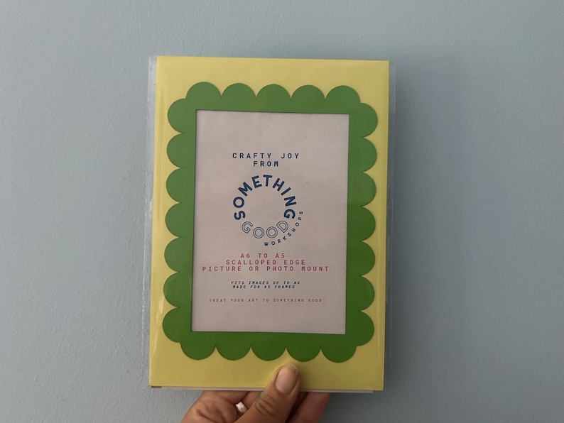 May include: A yellow picture frame with a green scalloped edge. The frame has a white interior with text that reads "Crafty Joy From Something Good Workshops". The frame is designed to hold A6 sized pictures or photo mounts.