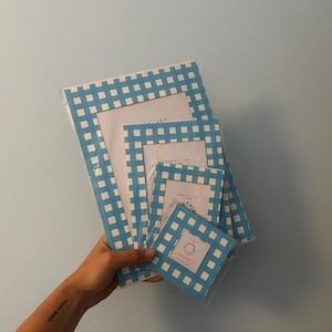 Op de afbeelding: Een stapel blauw en wit geruite fotolijsten in verschillende maten. De lijsten worden in een hand gehouden tegen een lichtblauwe achtergrond. De lijsten hebben de tekst "Crafts Joy From Esthia".
