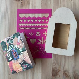 May include: A craft kit with a book titled "SOMETHING GOOD" and a pink stencil with decorative cutouts. A wooden frame with a copper-colored insert is also present. The items are arranged on a light wood surface.