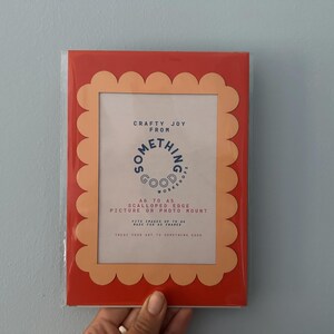 May include: A red picture frame with a scalloped edge and a peach-colored interior. The frame contains text that reads "Crafty Joy From Something Good Workshop" and "As To As Scalloped Edge Picture or Photo Mount."