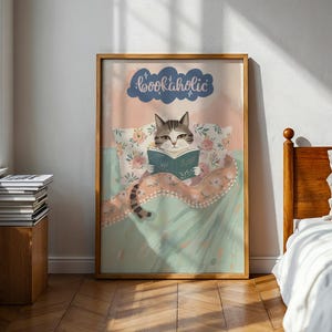以下が含まれることがあります： ベッドで本を読む猫が描かれた、額入りのアートプリントです。「bookaholic」の文字が猫の上にあります。イラストはパステルカラーを使用しています。猫はグレーと白で、寝具には花柄があります。