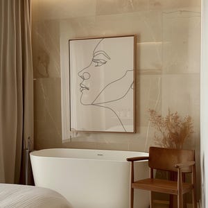 Könnte beinhalten: Ein modernes Badezimmer mit einer freistehenden weißen Badewanne, einem Holzstuhl und einem gerahmten Linienporträt eines Gesichts. Die Wände sind in einem hellbeigen Farbton gefliest, und der Raum wird von sanftem Licht erhellt.