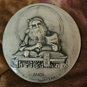 May include: A round, gray ceramic Christmas plate featuring a detailed relief of Santa Claus reading a book. The word "Christmas" is prominently displayed, along with the text "SANTA" and the date "December 4, 1920".
