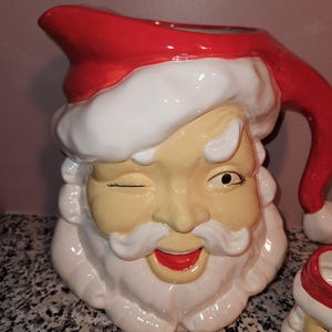 Può includere: Una brocca di Babbo Natale in ceramica con cappello e manico rossi, barba bianca ed espressione ammiccante. La brocca ha un viso color crema e una bocca rossa. Il design è festoso e adatto alle decorazioni natalizie.