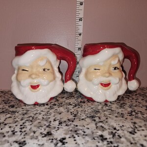 Può includere: Due tazze in ceramica di Babbo Natale. Ogni tazza presenta un volto di Babbo Natale che ammicca, con un cappello rosso e barba bianca. Le tazze sono alte circa 10 cm.