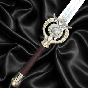 Puede incluir: Una espada con hoja de plata metálica y empuñadura envuelta en cuero marrón. La guarda es de metal dorado con un diseño circular y una piedra texturizada en el centro, rematada con un pomo a juego en tono dorado.